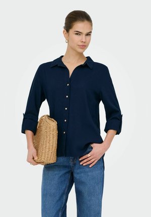 Donna con capelli raccolti indossa una camicia blu navy con bottoni e jeans blu, tiene una borsa intrecciata color tan al fianco e posa con una mano in tasca.