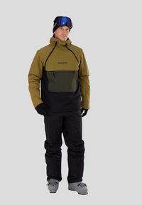 Sněžná bunda v olivově zelené a černé barvě s přední kapsou na zip, kapucí a texturovanou tkaninou. V kombinaci s černými sněhovými kalhotami a šedými lyžařskými botami.