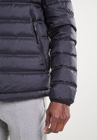 Giubbotto imbottito in blu scuro, caratterizzato da un design trapuntato, tasca laterale con zip e polsini elasticizzati per una vestibilità sicura. Tessuto liscio e lucido.