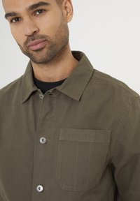 Olivenfarvet bomulds- skjortejakke med knaplukning, spidse kraver og en brystlomme. Har metal knapper og kontrastsyninger.