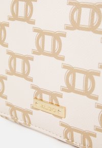 Beige purse with embossed logo pattern in gold. Features a metallic logo plate and textured surface. Stitched edges complement the design.