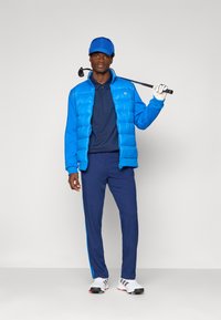 Homem em boné azul de golfe, casaco acolchoado, polo azul-marinho e calças, segurando um taco de golfe sobre o ombro, usando luva branca e sapatos de golfe.
