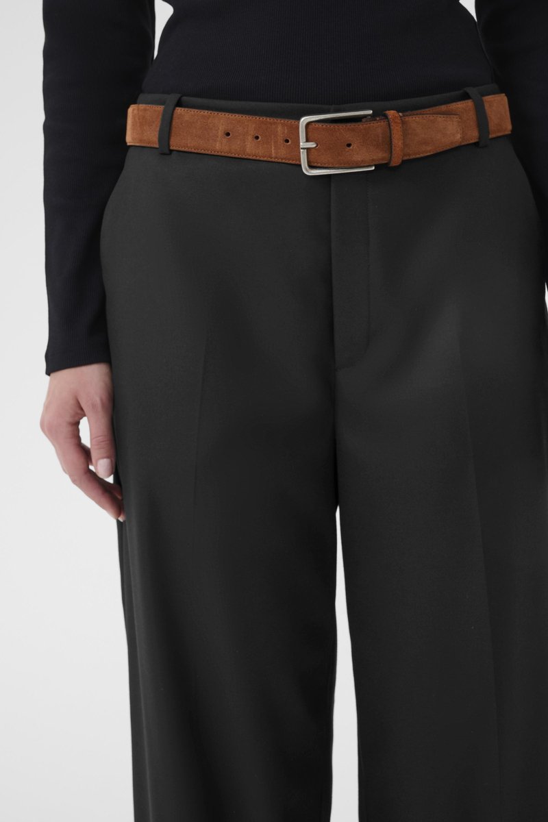 Pantalon noir en tissu lisse, à taille haute. Une ceinture en daim marron apporte un contraste avec une boucle en métal argenté.
