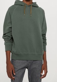 Felpa verde con cappuccio regolabile, polsini e orlo a coste. Tessuto morbido, presenta cuciture a contrasto e una piccola etichetta con logo in vita.