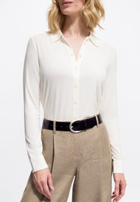 Femme portant une chemise crème à manches longues et boutons rentrée dans un pantalon beige taille haute avec une ceinture noire et une boucle en argent.