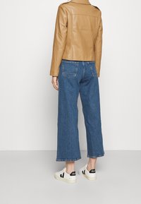 Une personne portant un jean bleu court, des baskets blanches avec un logo noir, et une veste en cuir beige, se tenant devant un fond blanc uni.