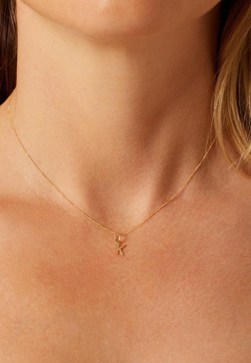 Goldene zarte Kettenhalskette mit einem kleinen Anhänger in Großbuchstaben "K", liegend auf einer glatten, gebräunten Hautoberfläche.