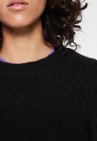 Schwarzer gerippter Pullover mit rundem Halsausschnitt; über einem lila Hemd getragen. Der Stoff wirkt weich und hat eine strukturierte Oberfläche.