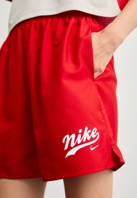 Rdeče atletske kratke hlače, narejene iz gladkega materiala, z elastičnim pasom in stranskimi žepi. Na levi strani je prikazana bela Nike logotip.