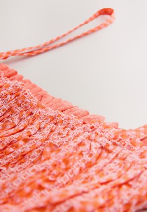 Coral pink dress with smocked fabric, ruffled edges, and thin straps. A checkered pattern accents part of the fabric, creating texture.