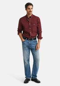 Man with mustache wearing red plaid button-up shirt, light blue jeans, black belt, and black shoes, standing with one hand in pocket.