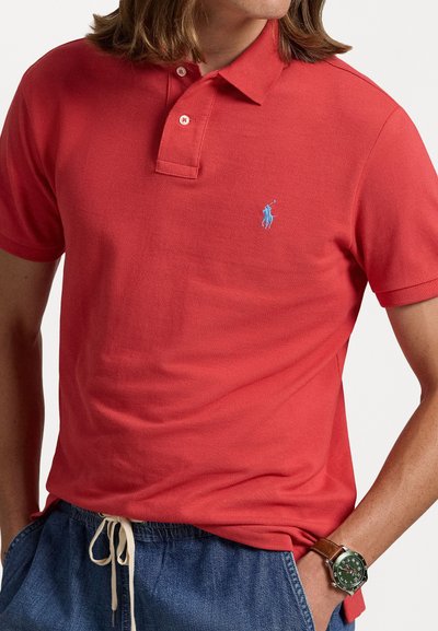 Röd polo tröja i texturerad bomull, med en treknappslucka och en liten blå logotyp på bröstet. Avslappnad stil med denimshorts.