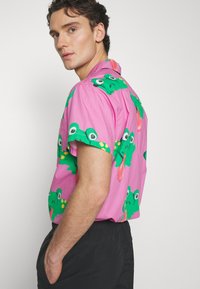 Camisa de manga curta rosa com padrões de sapos verdes, apresentando grandes olhos e línguas. Tecido leve com um corte descontraído.
