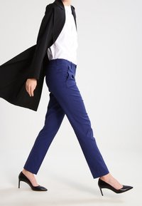 Blå mönstrade byxor med skräddarsydd passform, i kombination med en vit skjorta och svart kappa, kompletterat med svarta högklackade skor.