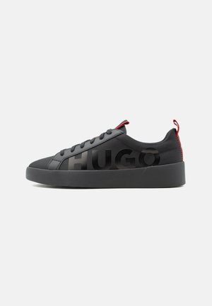 Black low-top sneaker with black sole, black laces, red pull tabs, and large black "HUGO" text on the side.