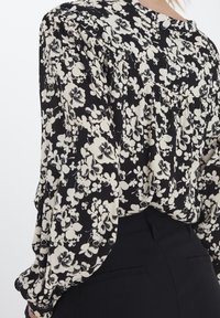 Černá blůza s bílým květinovým vzorem, kulatým výstřihem a nabíraným detailem na zadní straně. Dlouhé rukávy mají elastické manžety.