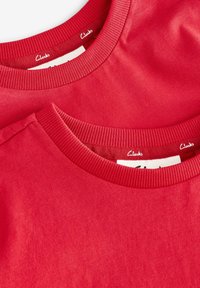 Twee rode T-shirts met geribbelde ronde hals, beide van katoen. Het ene shirt is over het andere gelegd, met een logo-label aan de binnenkant van de kraag.