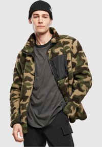 Vihreä- ja ruskeasävyinen maastokuvioinen fleecetakki, jossa on musta vetoketjulla suljettava tasku. Päällä harmaa paita. Takissa on korkeahko kaulus ja teksturoitu kangas.