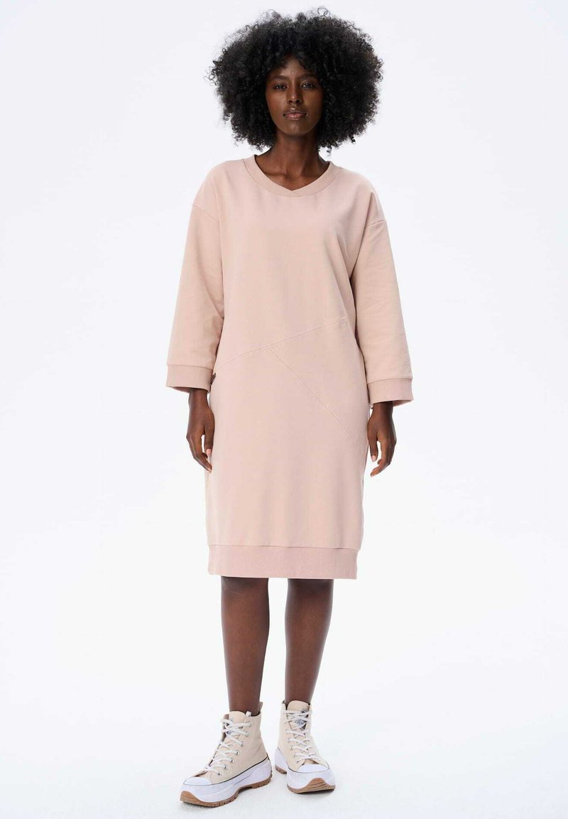 Leicht rosa Sweatkleid in lockerer Passform, mit langen Ärmeln, einem V-Ausschnitt und asymmetrischem Design, kombiniert mit beigen High-Top-Sneakers.