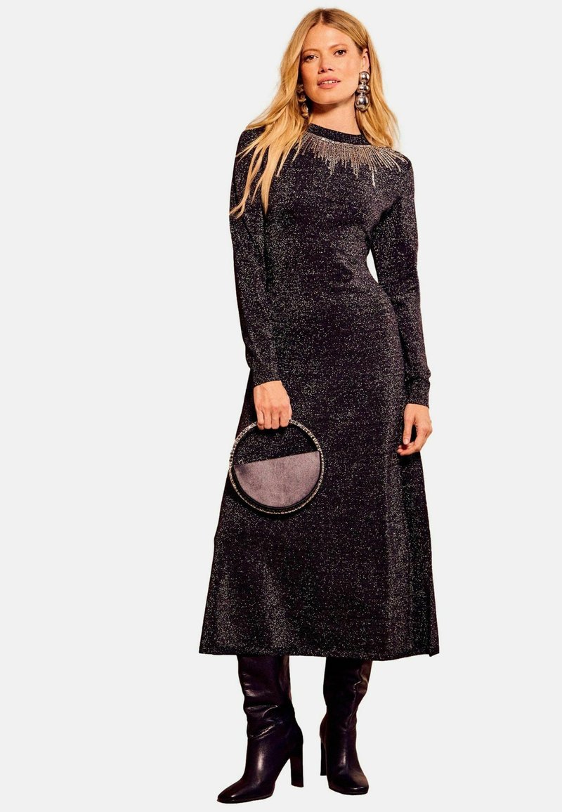 Abito lungo nero scintillante con silhouette aderente, scollatura rotonda e dettagli di perline. Abbinato a una borsa circolare e stivali fino al ginocchio.