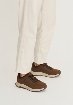 Persoon die beige broek en bruine vetersneakers met dikke witte zolen draagt tegen een lichte achtergrond.