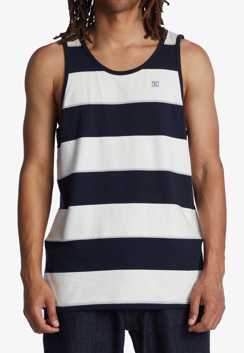 Homme portant un débardeur sans manches à larges rayures horizontales bleu marine et blanches, debout contre un fond blanc.