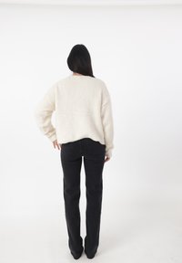 Maglione di tessuto cremoso con spalle scese abbinato a pantaloni neri a zampa, visto da dietro su uno sfondo bianco.