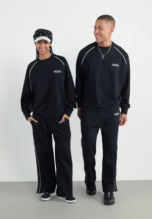 Felpe e pantaloni neri con accenti bianchi, con un logo sul petto, realizzati in tessuto morbido. Vestibilità comoda, entrambi i modelli indossano sneaker.