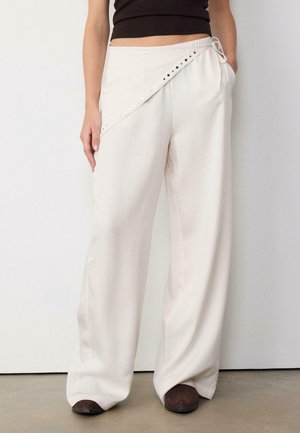 Kvinde iført hvide bukser med vidde og asymmetrisk klap med sølvnitter, sort kort top og brune sko, stående op ad en hvid væg.