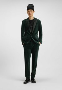 Dunkelgrüner Samtanzug mit schmal geschnittenem Sakko, satinierten Revers und passenden Hosen. Darunter ein schwarzes Rundhals-Shirt und eine schwarze Mütze.