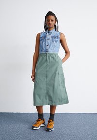 Jachetă din denim, la nivelul taliei, cu guler, asortată cu o fustă verde de lungime medie, care are un buzunar lateral. Tenis portocalii și negri completează ținuta.