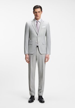 Mand iført lysegråt skræddersyet jakkesæt med to-knaps jakke, mønstret slips, hvid skjorte og sorte pæne sko, stående mod en hvid baggrund.