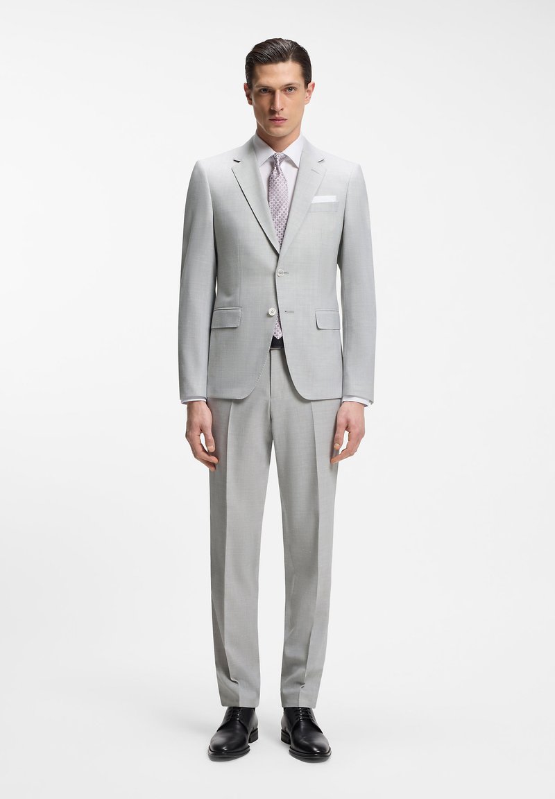 Άνδρας με ανοιχτό γκρι ραμμένο κοστούμι με σακάκι δύο κουμπιών, γραβατα με μοτίβο, λευκό πουκάμισο, μαύρα παπούτσια, στέκεται μπροστά σε λευκό φόντο.