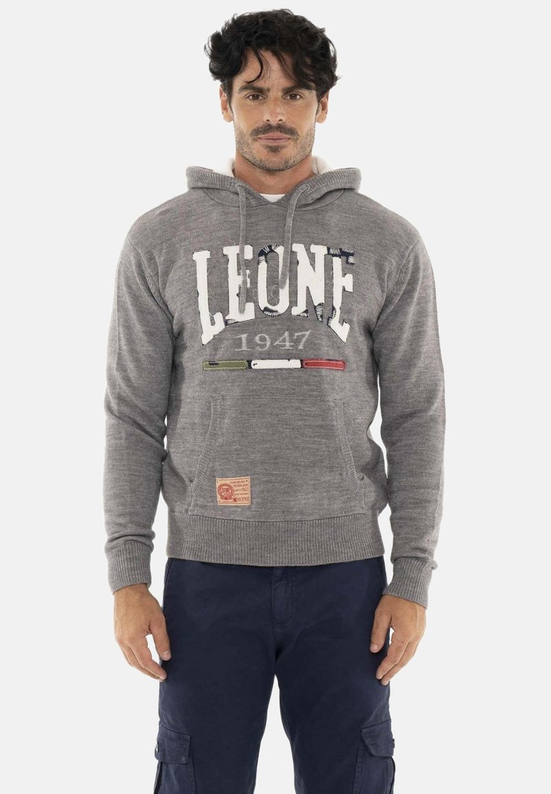 Homme aux cheveux foncés portant un sweat à capuche gris avec le texte « LEONE 1947 » et les couleurs du drapeau italien, associé à un pantalon cargo bleu marine, debout, droit.