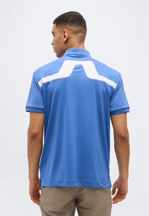 Mees, kes kannab sinist lühikeste varrukatega polosärki, millel on valge geomeetriline muster ülakehal selja peal, seisab ühevärvilise tausta ees.