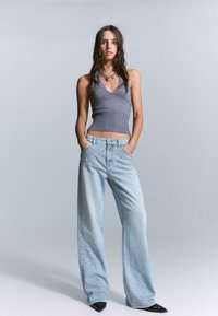 Light blue wide-leg jeans with studded accents on the sides, paired with a gray ribbed sleeveless top. Model poses with hands in pockets.