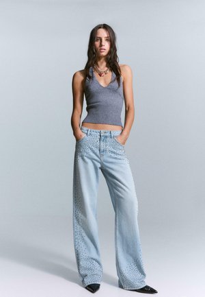 Light blue wide-leg jeans with studded accents on the sides, paired with a gray ribbed sleeveless top. Model poses with hands in pockets.