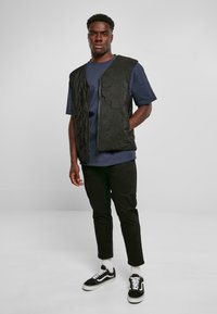 Zwarte gevoerde vest met ritssluiting, gedragen over een marineblauw t-shirt. Model heeft het gecombineerd met zwarte broek en zwarte sneakers met witte zolen.