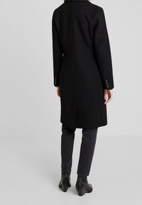 Schwarzer Wollmantel mit maßgeschneiderter Silhouette, Reverskragen und geknöpften Manschetten, kombiniert mit dunklen Jeans und schwarzen Stiefeletten.