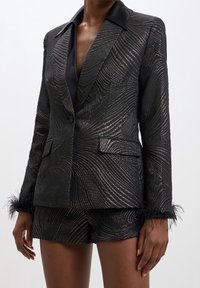 Schwarzer Blazer mit glänzendem, strukturiertem Muster, Satin-Revers und Federakzenten an den Ärmelabschlüssen, kombiniert mit passenden Shorts.