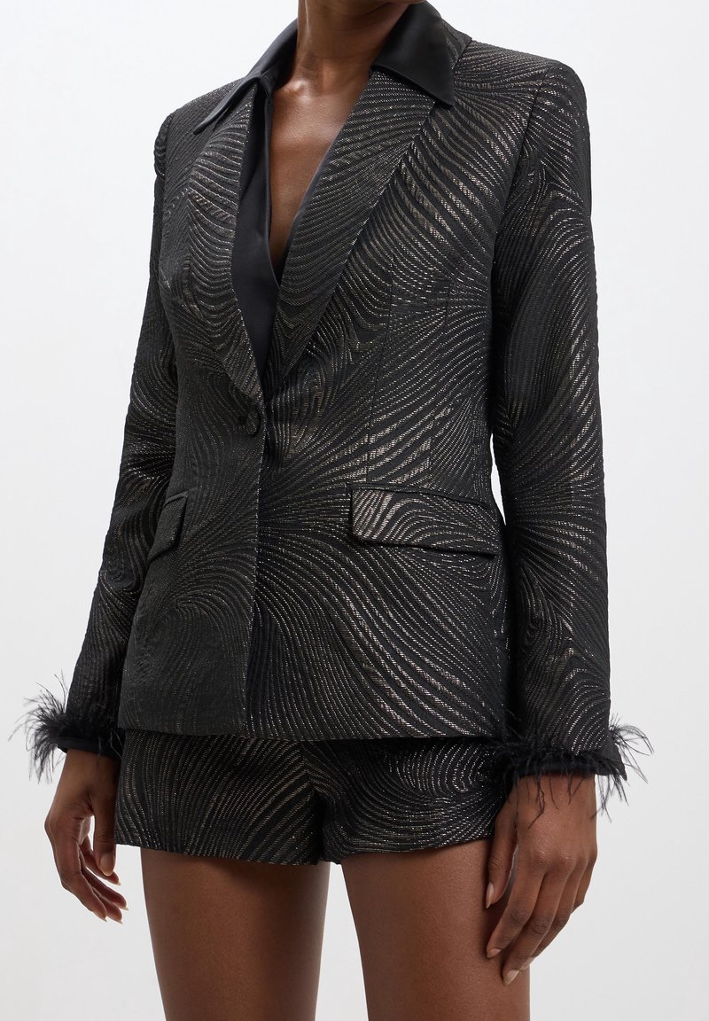 Schwarzer Blazer mit glänzendem, strukturiertem Muster, Satin-Revers und Federakzenten an den Ärmelabschlüssen, kombiniert mit passenden Shorts.