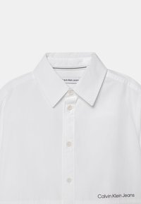 Biała koszula z długim rękawem z kołnierzykiem, przednim zapięciem na guziki oraz subtelnym logo near na dolnej krawędzi. Gładka faktura materiału.