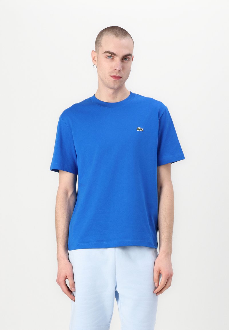 Μπλε βαμβακερό μπλουζάκι με στρογγυλή λαιμόκοψη, κοντά μανίκια και μικρό πράσινο λογότυπο στο αριστερό στήθος. Απλός σχεδιασμός με casual εφαρμογή.