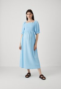 Rochie albastru deschis, până la genunchi, cu bust smockat, decolteu pătrat și mâneci bufante. Fabricată dintr-un material texturat, asortată cu sandale maro.