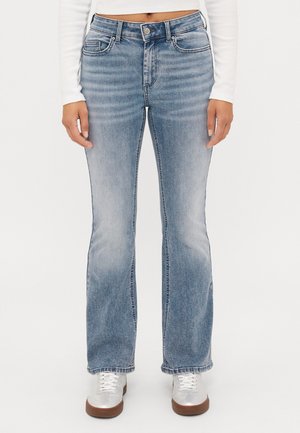 Frau trägt hellblaue ausgebleichte Bootcut-Jeans, weißen langärmligen Cropped-Top und weiße Sneakers mit braunen Sohlen, steht vor weißem Hintergrund.