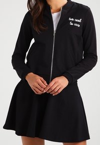 Vrouw in een zwarte zip-up jas met de witte tekst "too cool to care", gecombineerd met een zwarte flare rok en een wit topje eronder.
