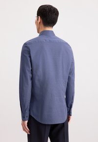 Uomo che indossa una camicia a maniche lunghe blu navy con pois bianchi, in piedi di spalle contro uno sfondo semplice.