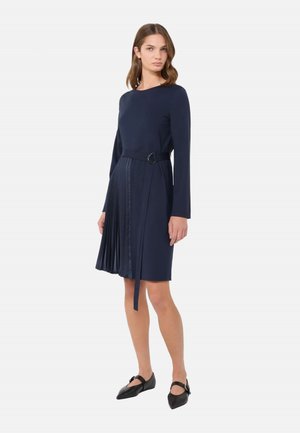 Donna che indossa un abito blu navy a maniche lunghe con dettaglio gonna plissettata e ballerine nere appuntite, in piedi su sfondo bianco semplice.