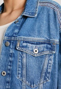 Jasnoniebieska kurtka dżinsowa, z wyprofilowanym kołnierzem, kieszenią na piersi, metalowymi guzikami i dekoracyjnym stębnowaniem na ramionach.