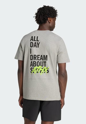 Man draagt een lichtgrijs T-shirt met zwarte en neongroene tekst op de rug met de tekst "All day I dream about ATFAS" en zwarte shorts.